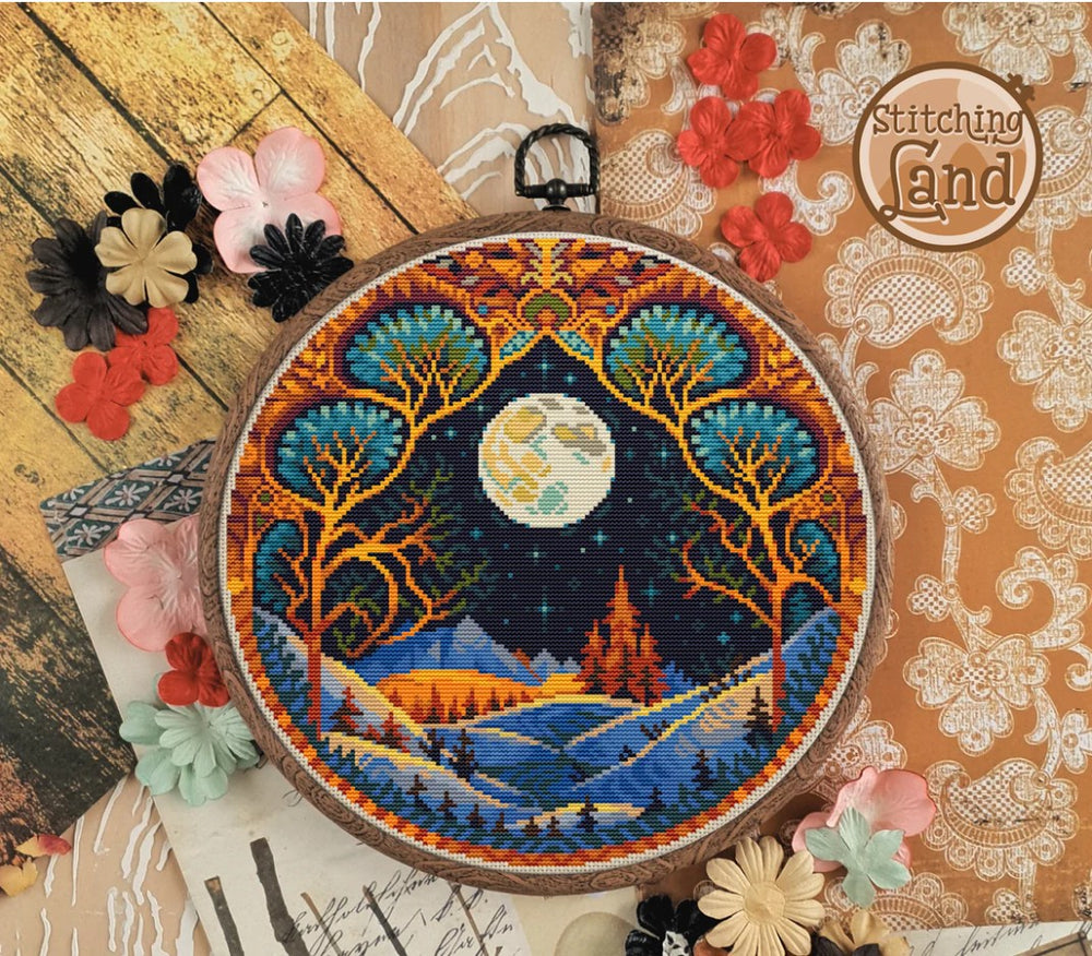 Stamped cross stitch design of landscape with trees and moon, surrounded by decorative flowers on a wooden surface.
