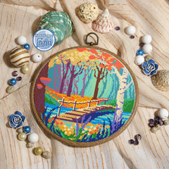Stamped cross stitch mockup of a Embroidery hoop with a colorful landscape design on a wooden surface with decorative items.