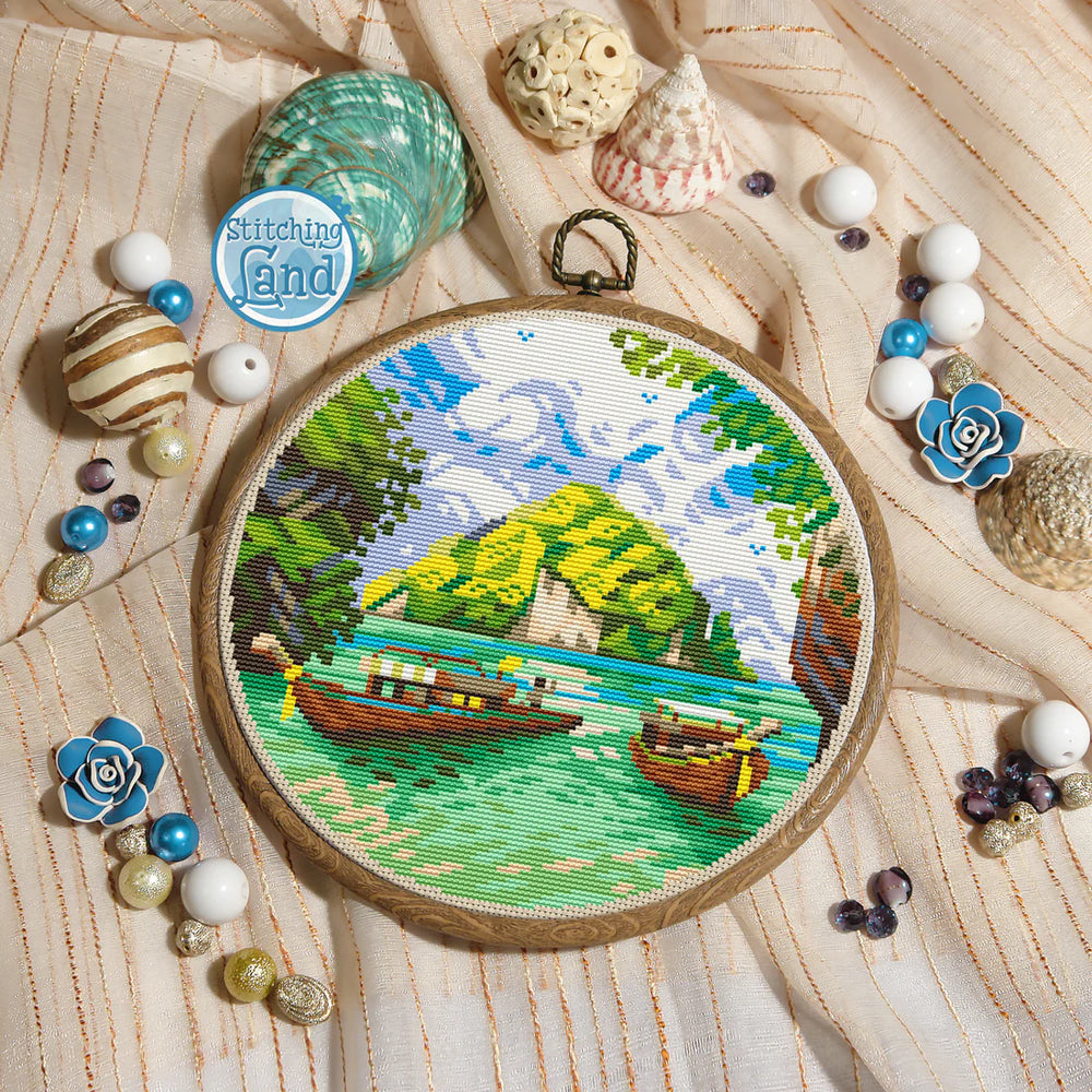 Stamped cross stitch mockup showing a Embroidery hoop with a scenic design surrounded by decorative shells and beads on a wooden surface.