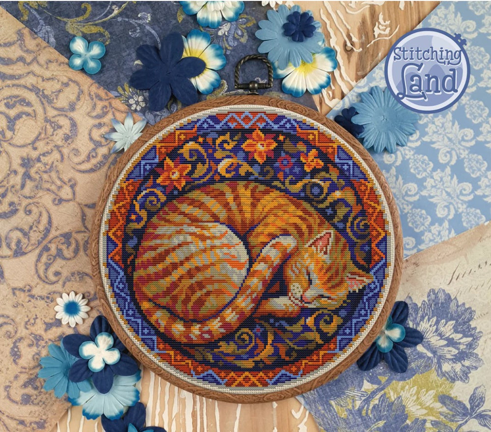 Embroidery hoop with a sleeping cat stamped cross stitch design on a decorative background with flowers and patterns.