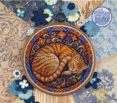 Embroidery hoop with a sleeping cat stamped cross stitch design on a decorative background with flowers and patterns.