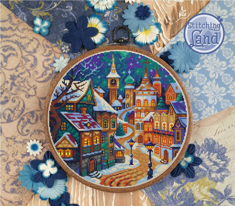 stamped cross-stitched village scene in an embroidery hoop with decorative flowers and 'Stitching Land' branding.