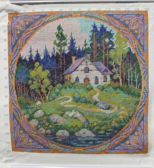 Cross stitch scene of a house surrounded by trees and nature, framed with decorative border.